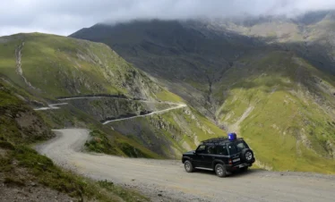 A 4x4 vehicle with roof rack on a gravel mountain road on the Abano Pass in Georgia, with serpentine switchbacks descending into a green valley below