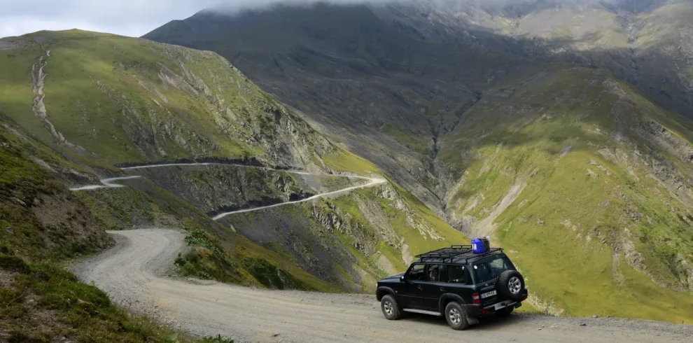 A 4x4 vehicle with roof rack on a gravel mountain road on the Abano Pass in Georgia, with serpentine switchbacks descending into a green valley below