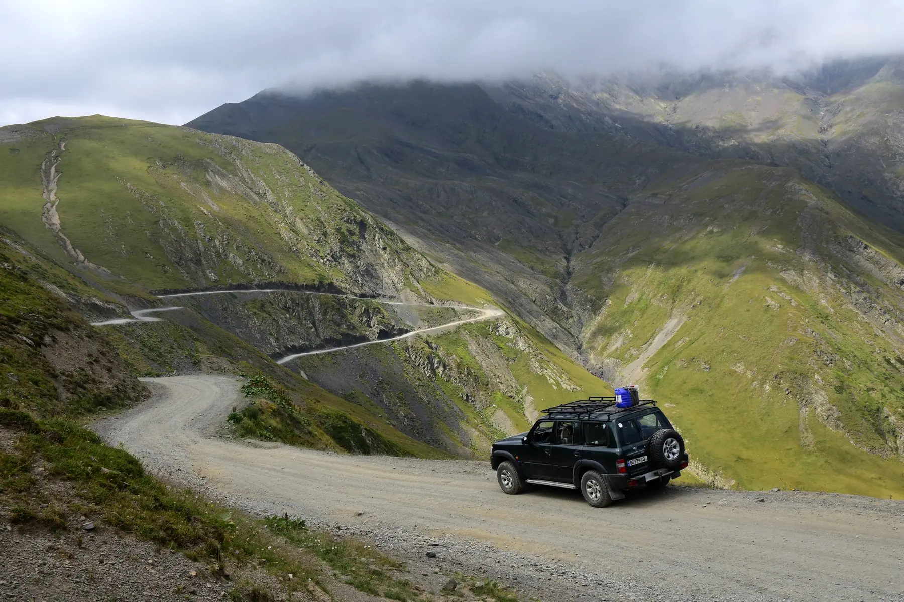 A 4x4 vehicle with roof rack on a gravel mountain road on the Abano Pass in Georgia, with serpentine switchbacks descending into a green valley below
