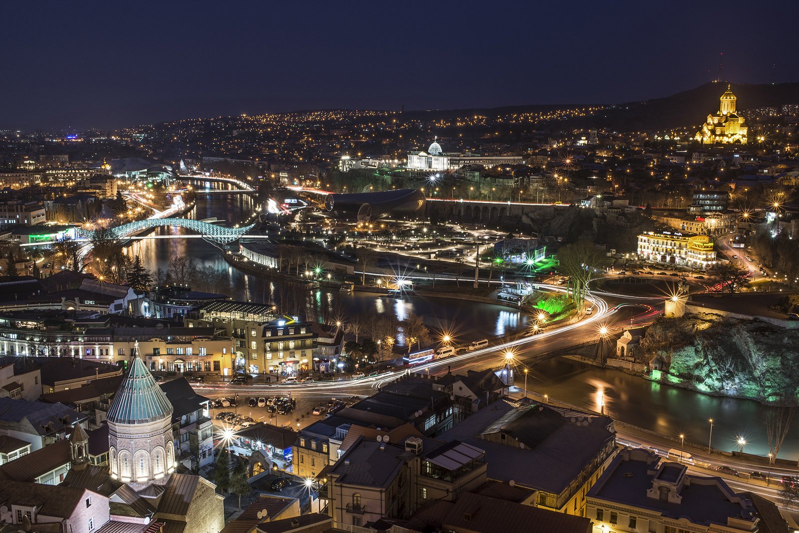 Night panorama of Tbilisi with the illuminated Mtkvari river, the Bridge of Peace and the Sameba Cathedral glowing on the hilltop, Georgia